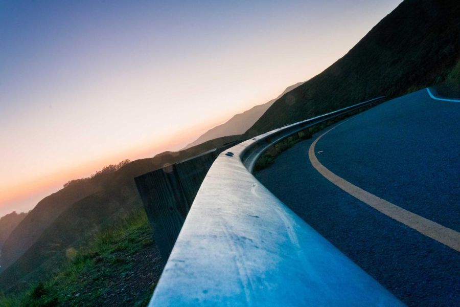 Guardrail curving along a mountain road at dusk, with hills fading into an orange sky