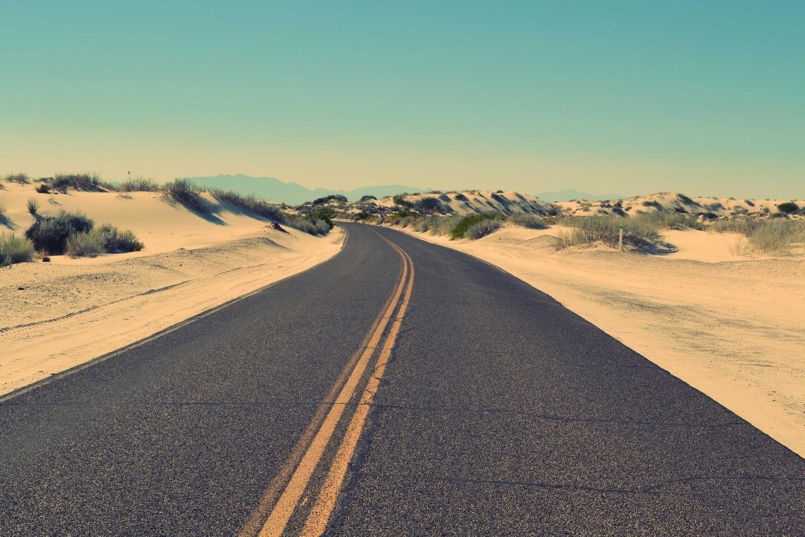 Two-lane desert road with a double yellow line curving between sand dunes